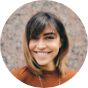Smiling woman with medium-length brown hair and bangs, wearing a rust-colored top.