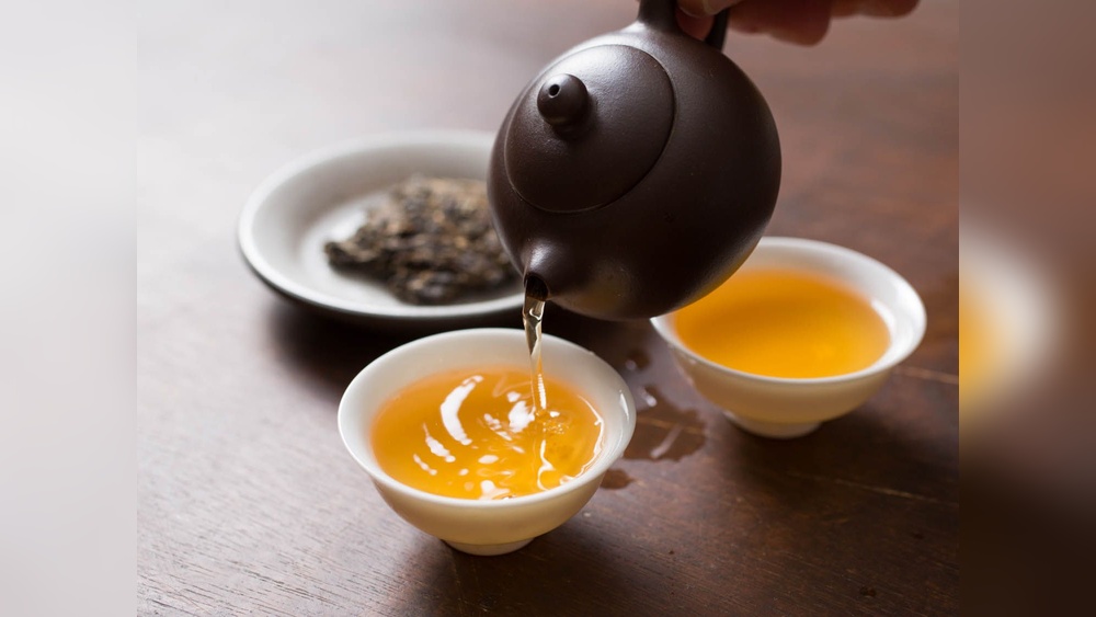 Pouring golden tea from a dark teapot into a white cup, with dried tea leaves in the background.