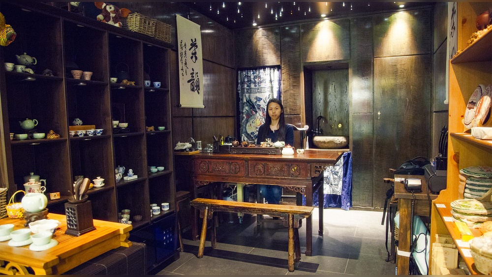 Woman serving tea in a traditional Chinese tea shop with shelves of teaware.