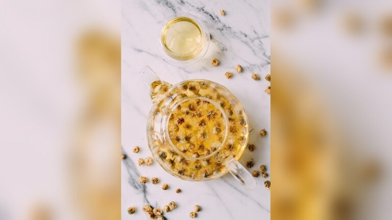 Glass teapot and cup filled with golden chrysanthemum tea on a marble surface.