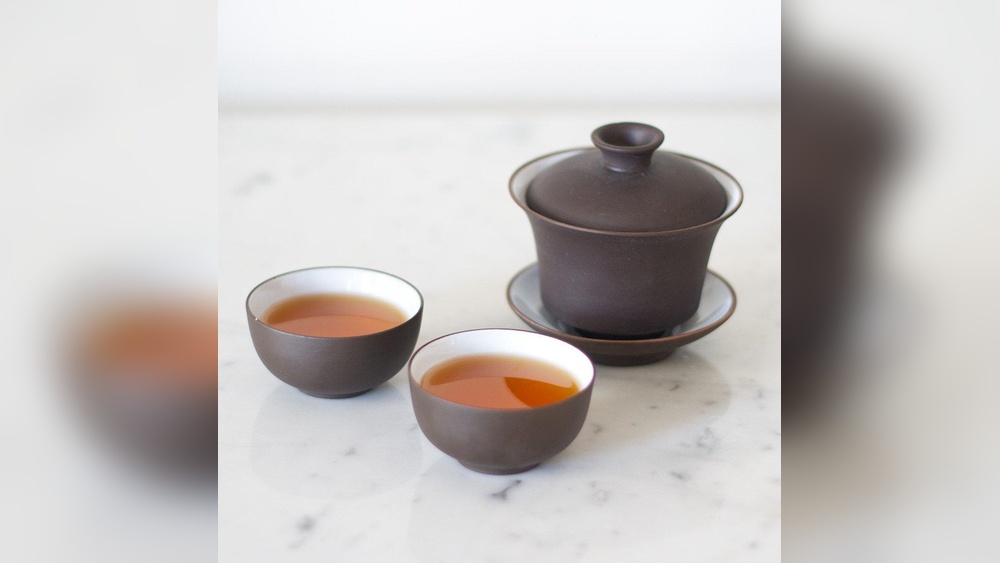 Brown ceramic gaiwan tea set with two cups filled with amber tea.